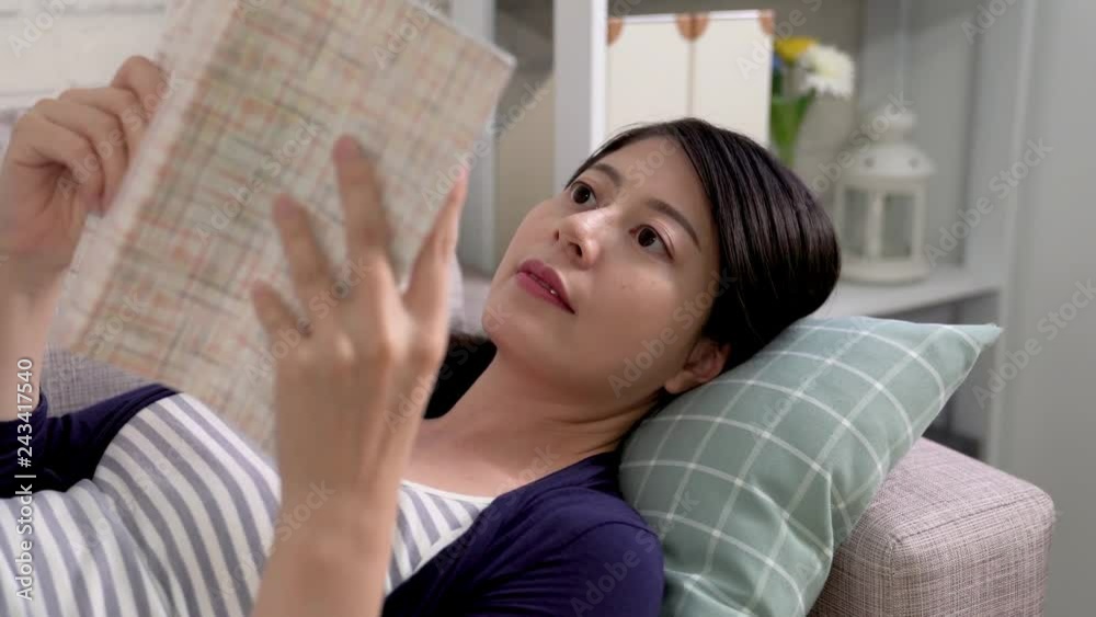 Young smiling woman sitting on sofa and looking up while reading romantic novel. beautiful asian woman thinking at home in a leisure time. Happy girl relaxing in bright living room in the morning. Young smiling woman sitting on sofa and looking up while reading romantic novel. beautiful asian woman thinking at home in a leisure time. Happy girl relaxing in bright living room in the morning.