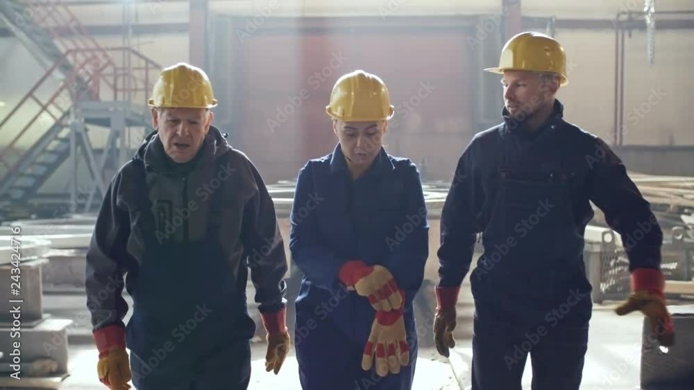 Dolly shot of female engineer in overalls, hard hat and gloves walking through metal fabrication facility and discussing work with male colleagues