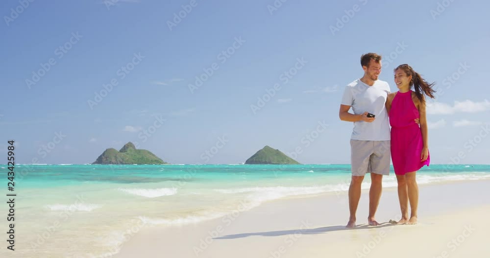 Beach couple using phone app smiling and laughing walking on beach having fun outside under the blue sky . Young Asian woman, Caucasian man. RED EPIC SLOW MOTION Beach couple using phone app smiling and laughing walking on beach having fun outside under the blue sky . Young Asian woman, Caucasian man. RED EPIC SLOW MOTION