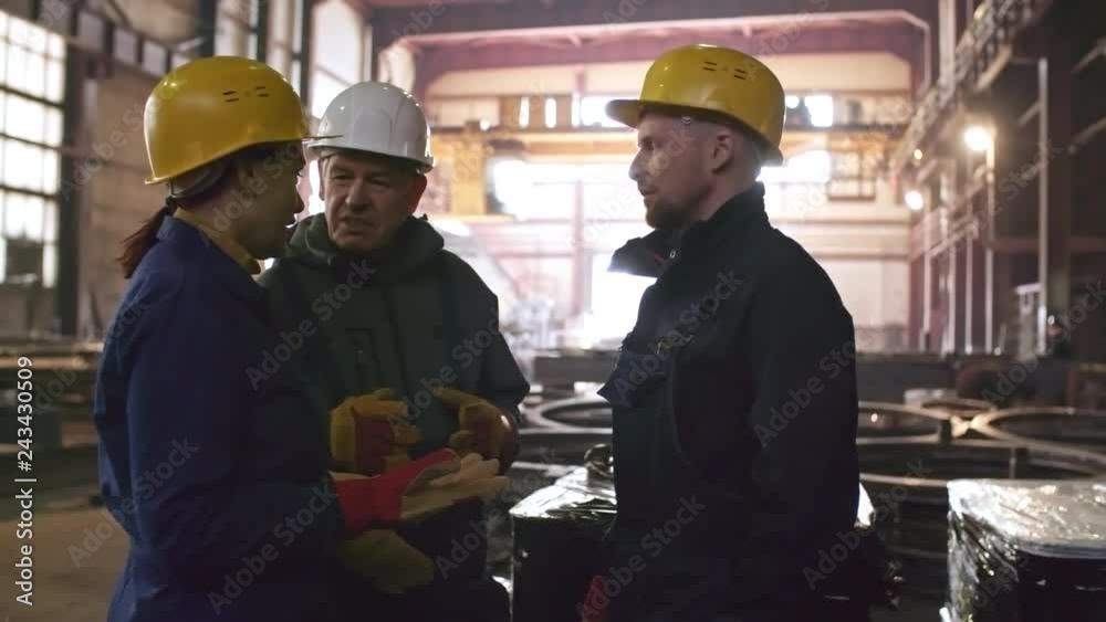 PAN shot of middle-aged female engineer in hard hat and gloves giving instructions to male workers at metal fabrication facility PAN shot of middle-aged female engineer in hard hat and gloves giving instructions to male workers at metal fabrication facility