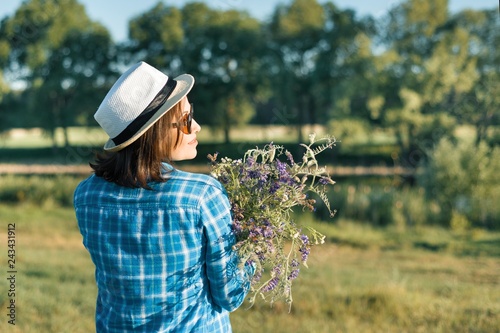 Outdoor summer portrait of woman with bouquet of wildflowers, straw hat. View from the back, nature background, rural landscape, green meadow, country style.