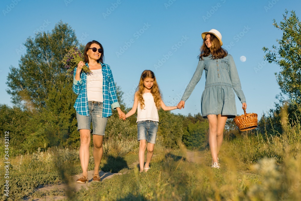 Happy mother and two daughters holding hands walking along rural country road with wildflowers, basket of berries. Sunny summer day, sunset, back view