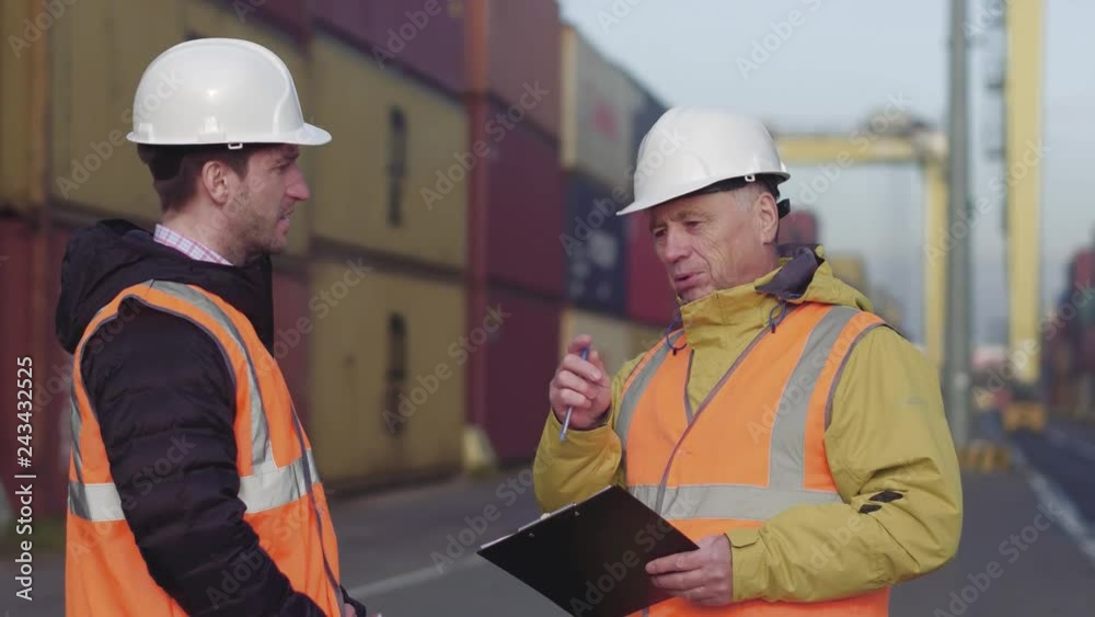 Port manager and a colleague tracking inventory while standing together by freight containers on a large commercial shipping dock Port manager and a colleague tracking inventory while standing together by freight containers on a large commercial shipping dock