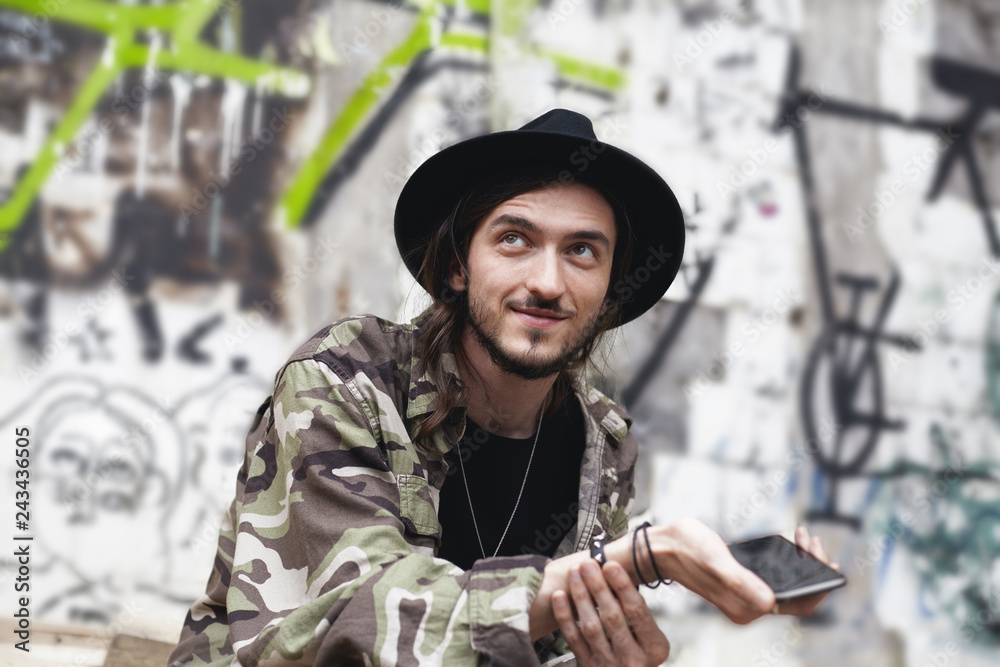 Style, fashion, online communication and modern technologies. Picture of fashionable cheerful bearded guy in good mood using electronic gadget, sitting against wall with street art and graffiti