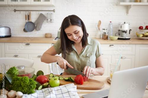 Charming brunette girl in her twenties learning how to make vegan lasagne  using sharp knife while cutting bell pepper on cooking board  siting in front of laptop  surrounded with all ingredients