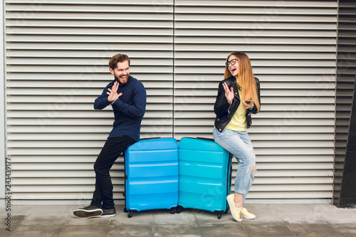 Cute couple is leaned on two suitcases on gray striped background. She has long hair, glasses,  yellow sweater, jacket, jeans. He wears black shirt, pants, beard. They are gratulating each other. photo