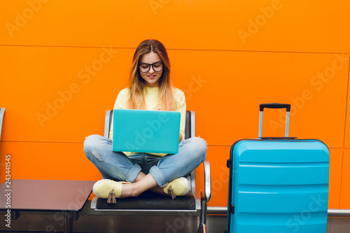 Young girl with long hair is sitting on chair on orange background. She wears yellow sweater and jeans. She is typing on laptop. photo