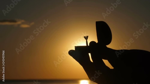 Close-up silhouette male hands opening engagement ring box with diamond proposal ring on bright orange sunset background. Marriage proposal with engagement ring to beloved woman in nature on seashore