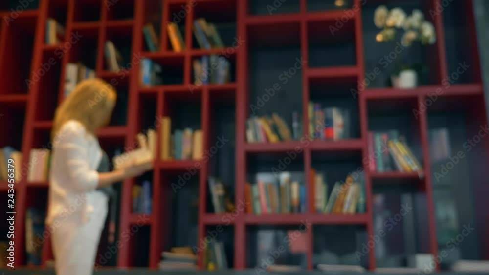Focusing on a mug with a hot drink, on a table in the library. Blur, Young woman, choosing a book to read, standing in front of a bookcase. Focusing on a mug with a hot drink, on a table in the library. Blur, Young woman, choosing a book to read, standing in front of a bookcase.