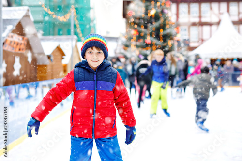 Happy little kid boy in colorful warm clothes skating on a rink of Christmas market or fair. Healthy child having fun on ice skate. Lot of people celebrating holiday and having active winter leisure