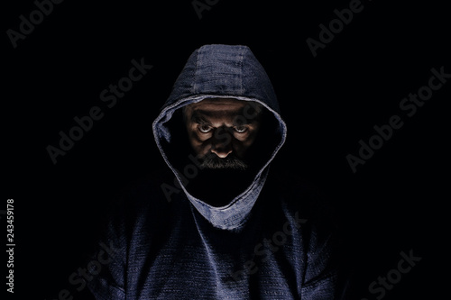Dark mysterious man with beard looking ominous, wearing blue hoodie, sitting in total dark