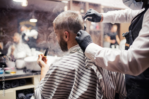 Hipster man client visiting haidresser and hairstylist in barber shop, smoking a pipe.