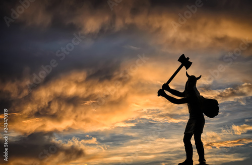 Dark silhouette of a Viking holding a double-sided axe against orange-blue stormy sunset sky with dark clouds, epic mythical illustration on Old Norse gods theme, free space on the left