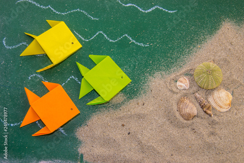 Photography Paper origami fish on board for writing with shells on the sand.