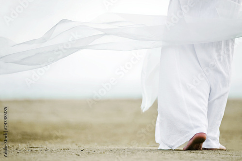 Photo of a man walking in a white robe on the sand. A picture from the back, a person is visible to the waist, clothes are fluttering in the wind