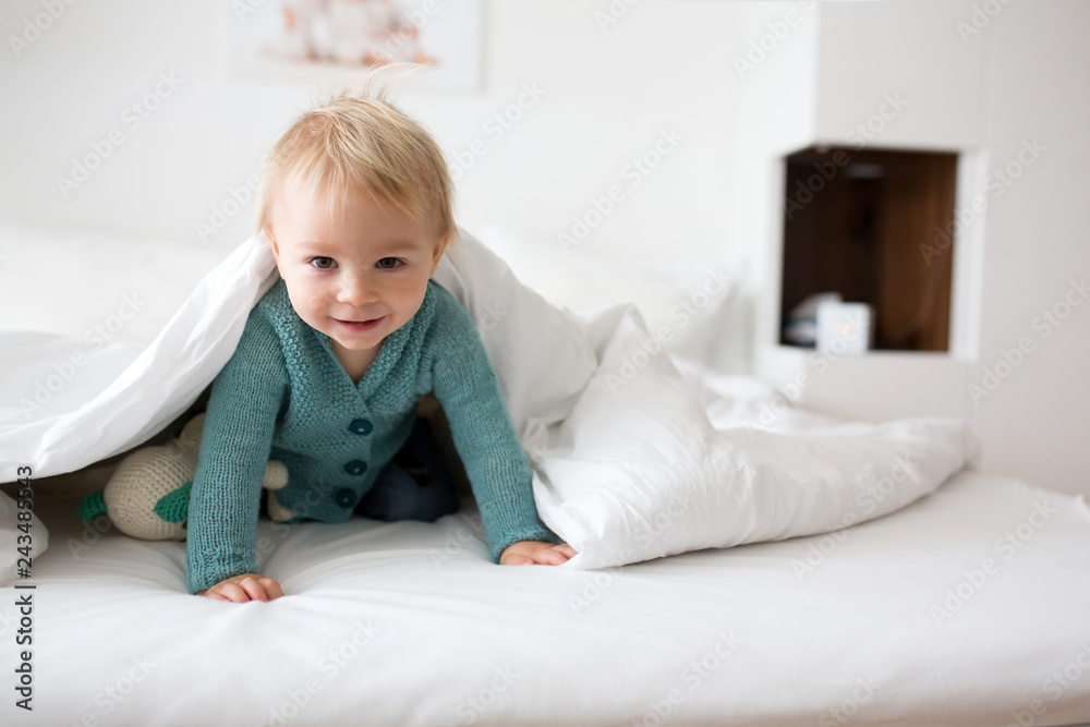 Little cute baby boy, child in knitted sweater, holding knitted toy, smiling happily at camera in white sunny, bright bedroom. Little cute baby boy, child in knitted sweater, holding knitted toy, smiling happily at camera in white sunny, bright bedroom.