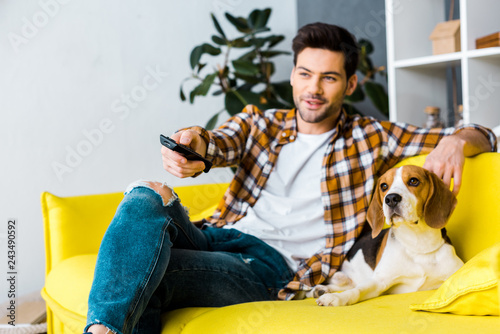 smiling man with remote control watching tv and sitting on sofa with dog