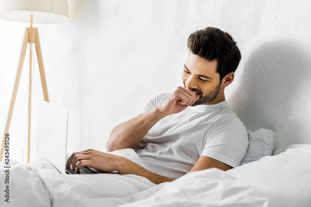 handsome laughing man using laptop in bed in the morning handsome laughing man using laptop in bed in the morning