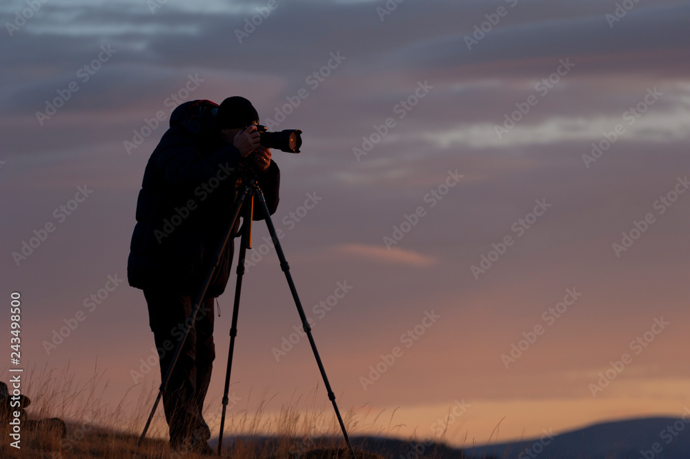 Photographer's silhouette early in the morning at sunrise carefully focusing and composing photos with his digital camera set on a tripod.