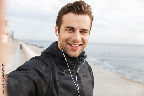 Image of smiling sportsman 30s in black sportswear and earphones, taking selfie photo on mobile phone while walking at seaside
