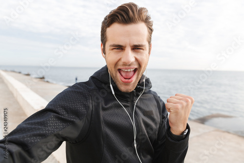 Image of cheerful sportsman 30s in black sportswear and earphones, taking selfie photo on mobile phone while walking at seaside