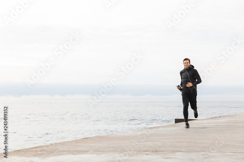 Image of excited sportsman 30s in black sportswear and earphones, working out and running by seaside