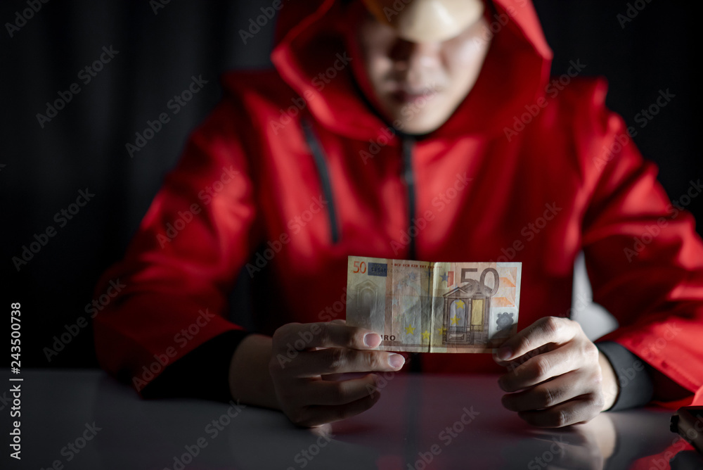 Mystery man wearing mask and red hoodie holding euro banknote on the table. Anonymous thief or robber committing a robbery. Crime and violence concept Mystery man wearing mask and red hoodie holding euro banknote on the table. Anonymous thief or robber committing a robbery. Crime and violence concept