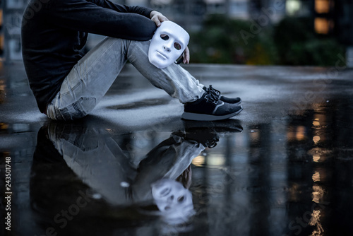 Reflection of mystery man with black jacket holding white mask sitting in the rain on rooftop of abandoned building. Bipolar disorder or Major depressive disorder. Depression concept