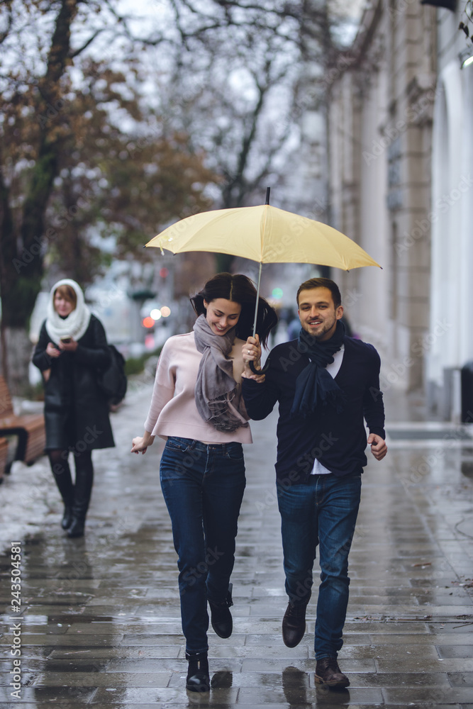 Fototapeta premium Beautiful couple, guy and his girlfriend dressed in casual clothes are running under the umbrella on the street in the rain.