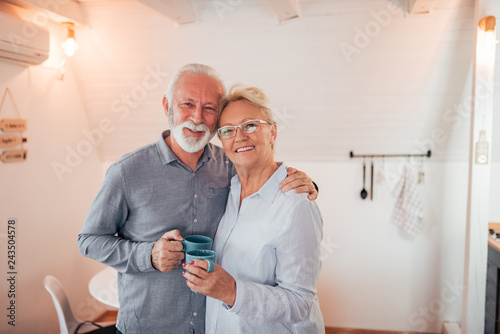 Home portrait of a senior couple holding mugs, looking at camera.