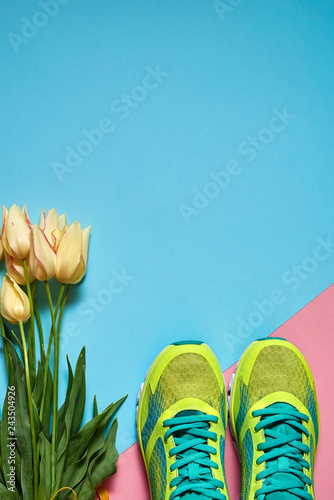 Pair of sport shoes and yellow tulips bouquet on colorful pastel background. New sneakers on pink and blue paper, copy space. Overhead shot of running foot wear. Top view, flat lay