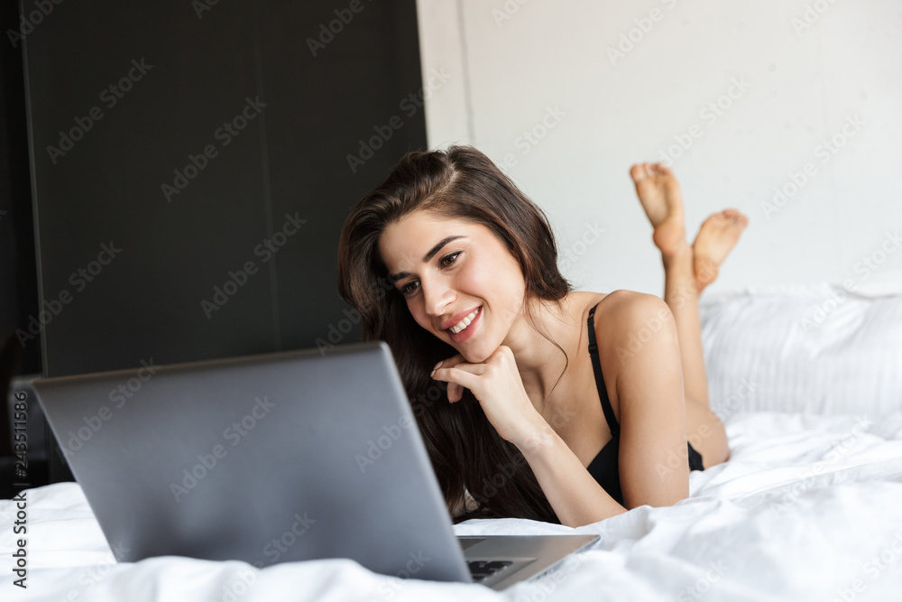 Fototapeta premium Beautiful brunette woman wearing lingerie using laptop computer lies in bed.