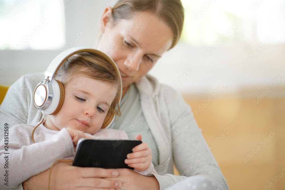 Mother and cute young daughter at home with cellphone and headphones