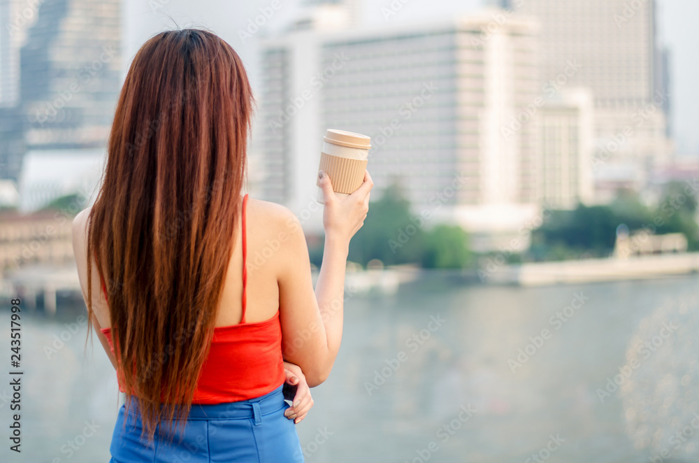 Rear view of a young woman standing at bangkok skyline with a cup of coffee, looking at the city after working in the evening.