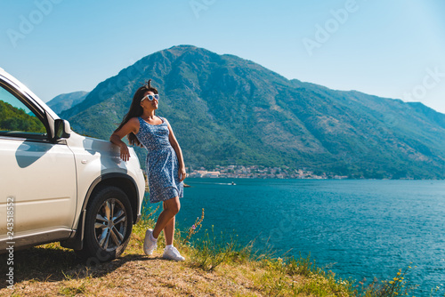 woman standing on cliff near white suv car with beautiful view of sea bay with mountains