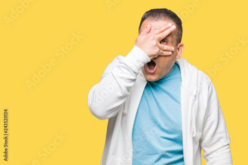 Middle age arab man wearing sweatshirt over isolated background peeking in shock covering face and eyes with hand, looking through fingers with embarrassed expression.