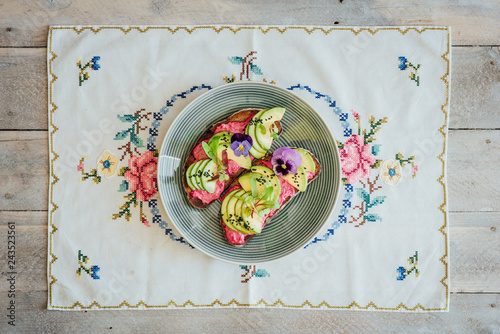 Top shot of two slices avocado rye toast on beetroot hummus with edible purple pansy flowers, micro greens and nigella seeds or onion seeds, on a blue plate, on embroided teatowel on white wooden back photo