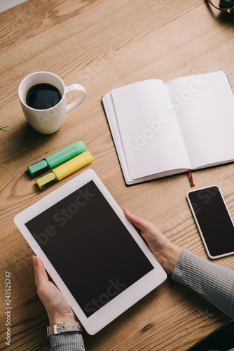 cropped view of woman holding digital tablet at table with smartphone, notebook and cup of coffee