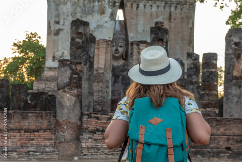 Travelling woman with backpack on Ancient sculpture of a standing Buddha in ruins of a Buddhist temple, historical park of Sukhothai, Thailand.