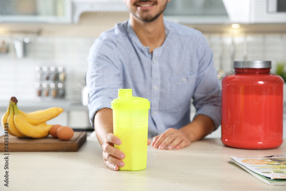 Young man holding bottle of protein shake at table with ingredients in kitchen Young man holding bottle of protein shake at table with ingredients in kitchen