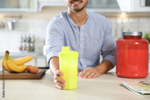 Young man holding bottle of protein shake at table with ingredients in kitchen
