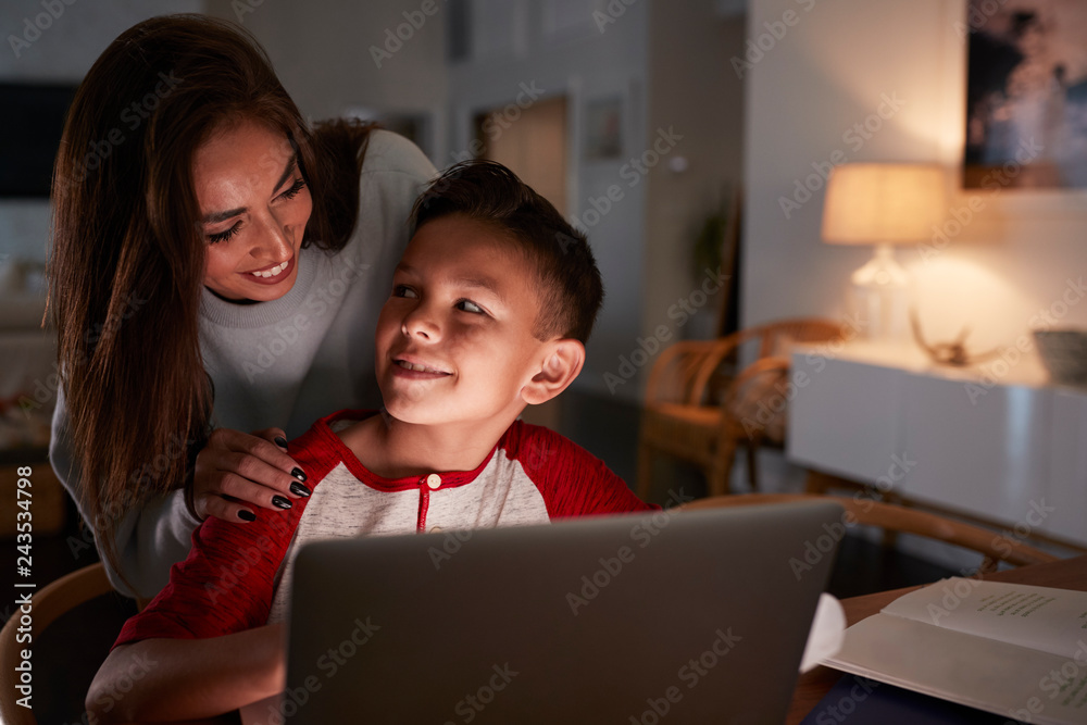 Hispanic woman looking over her sonÕs shoulder while he does his homework using laptop computer Hispanic woman looking over her sonÕs shoulder while he does his homework using laptop computer