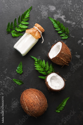 Coconut and milk in a bottle on a dark stone background decorated with fern leaves. Top view, flat lay.