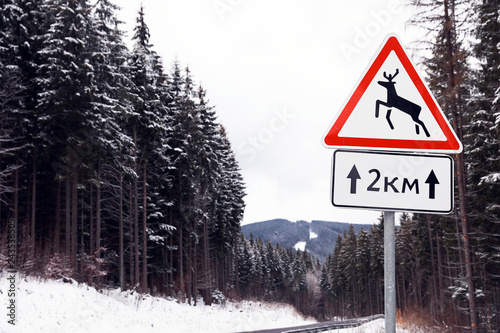 Canvas Print Wild animals warning traffic sign near road with snow on sides through forest