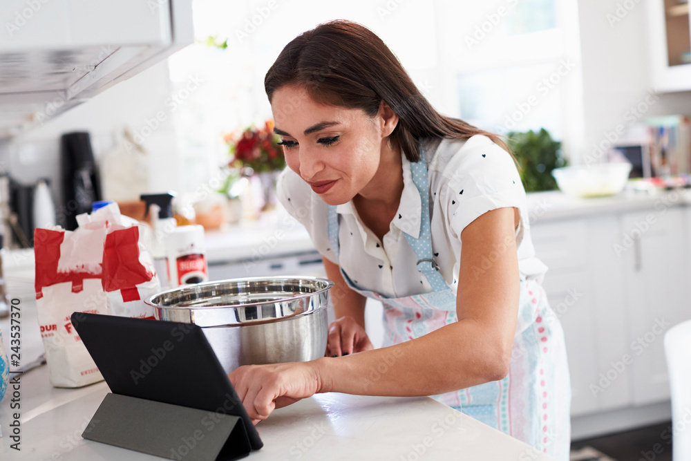 Millennial Hispanic woman preparing cake mixture checking the recipe on her tablet computer Millennial Hispanic woman preparing cake mixture checking the recipe on her tablet computer