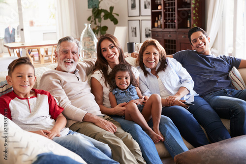Three generation Hispanic family sitting on the sofa watching TV, looking to camera