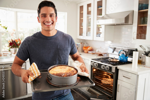 Smiling millennial Hispanic man standing in kitchen presenting the cake he has baked to camera photo