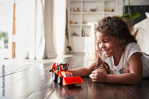 Young Hispanic girl lying on the floor in the sitting room playing with a toy digger truck
