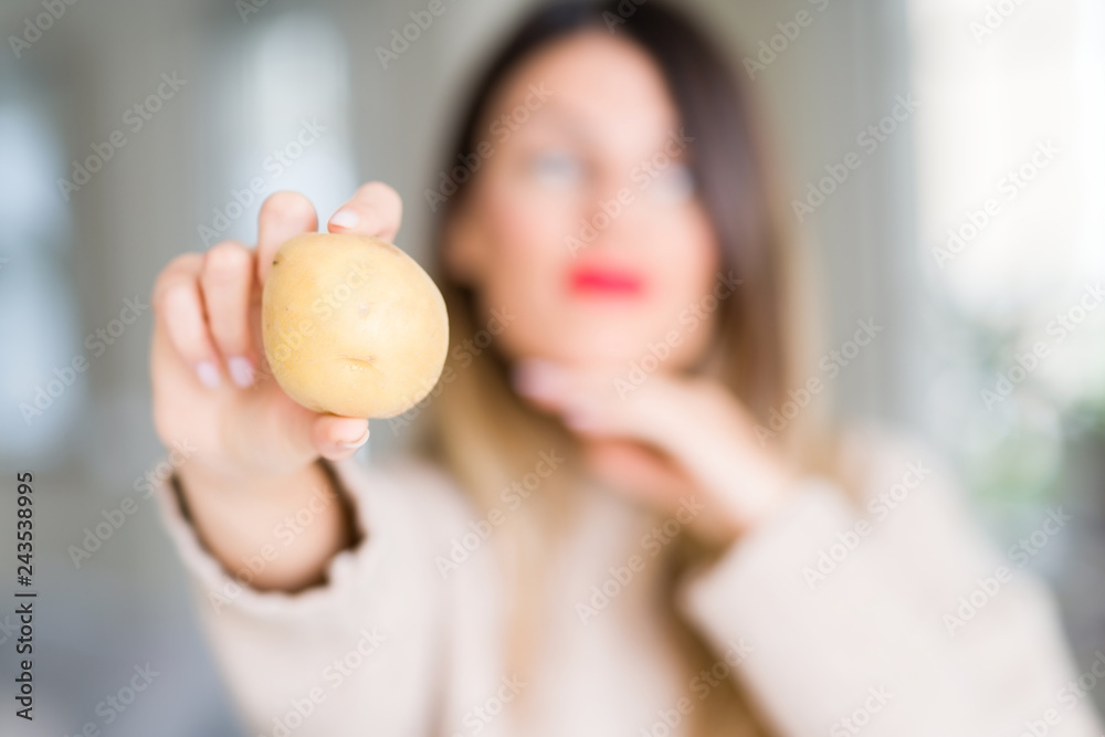 Young beautiful woman holding fresh potato at home serious face thinking about question, very confused idea Young beautiful woman holding fresh potato at home serious face thinking about question, very confused idea