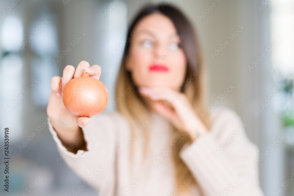 Young beautiful woman holding fresh onion at home serious face thinking about question, very confused idea Young beautiful woman holding fresh onion at home serious face thinking about question, very confused idea